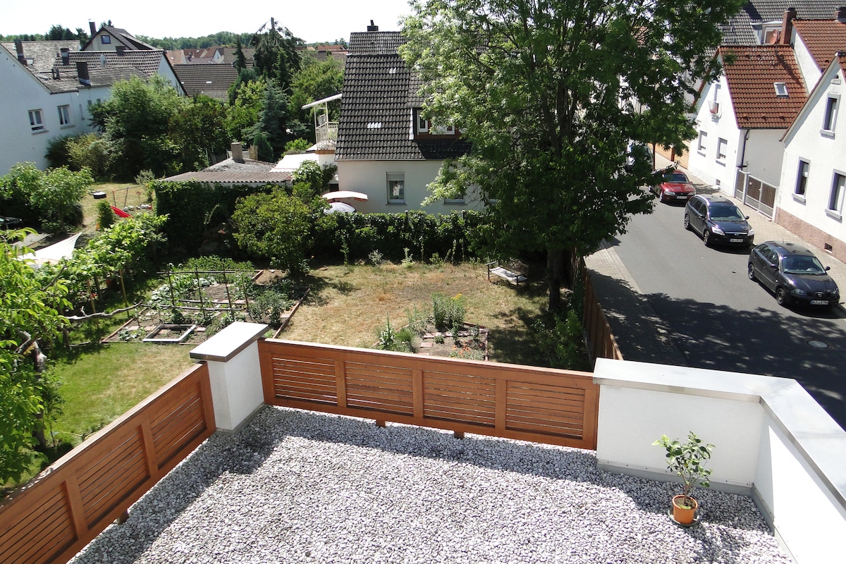 A view from a balcony overlooks a landscaped garden and neighboring houses. The garden features a neatly arranged plot with greenery, while a gravel area is visible on the balcony. A street runs adjacent to the property, lined with trees and residential buildings.