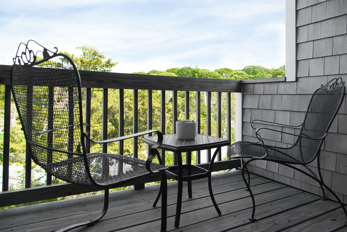 A small outdoor balcony features two metal chairs and a round table. A white planter sits on the table, surrounded by a railing that overlooks lush greens and trees in the distance, with a clear sky providing a serene backdrop.