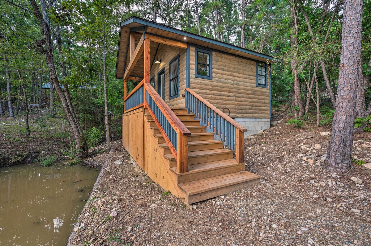 A cabin sits nestled among trees near a private pond. Sturdy wooden steps lead up to a welcoming entrance. The exterior is characterized by wood paneling and a blue trim around the windows, enhancing its rustic charm.
