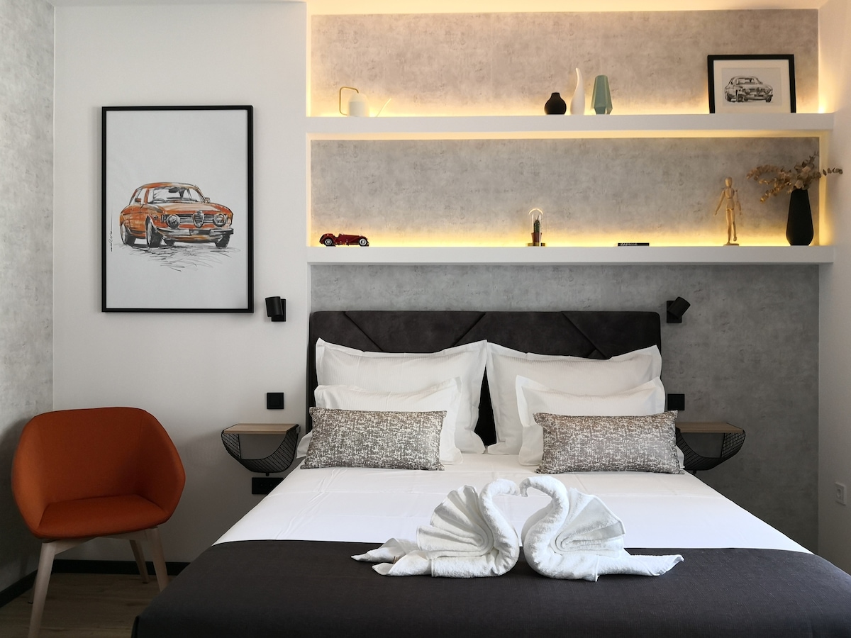 A comfortable Queen size bed is centered in the room, adorned with decorative pillows. A piece of artwork featuring a vintage car hangs above. Ambient lighting highlights the shelving above, which holds decorative items. An orange chair adds a pop of color against the neutral tones.