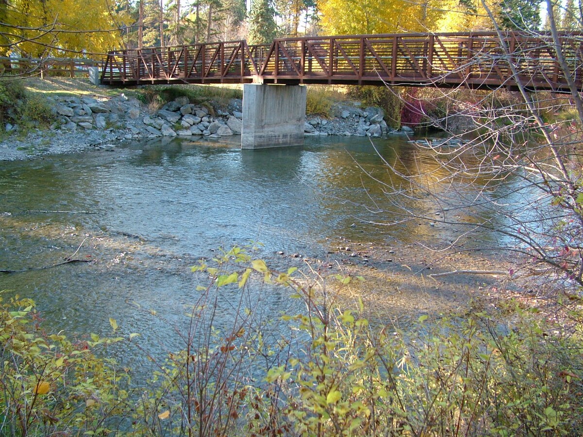 A wooden bridge crosses a gently flowing river, surrounded by rocks and autumn foliage. Colorful leaves line the banks, highlighting the tranquil setting. The water reflects the surrounding trees, creating a serene natural landscape.