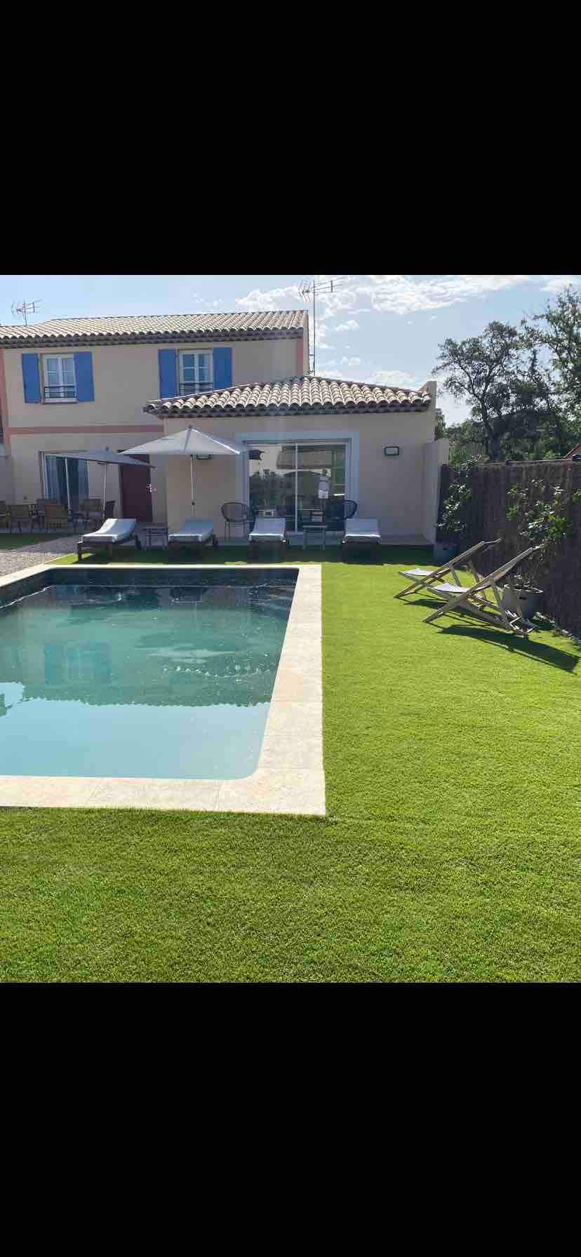 A private swimming pool is framed by manicured green grass, creating a relaxing outdoor space. Two lounge chairs are positioned nearby, while the house with blue-shuttered windows is visible in the background, bordered by trees and a clear blue sky.