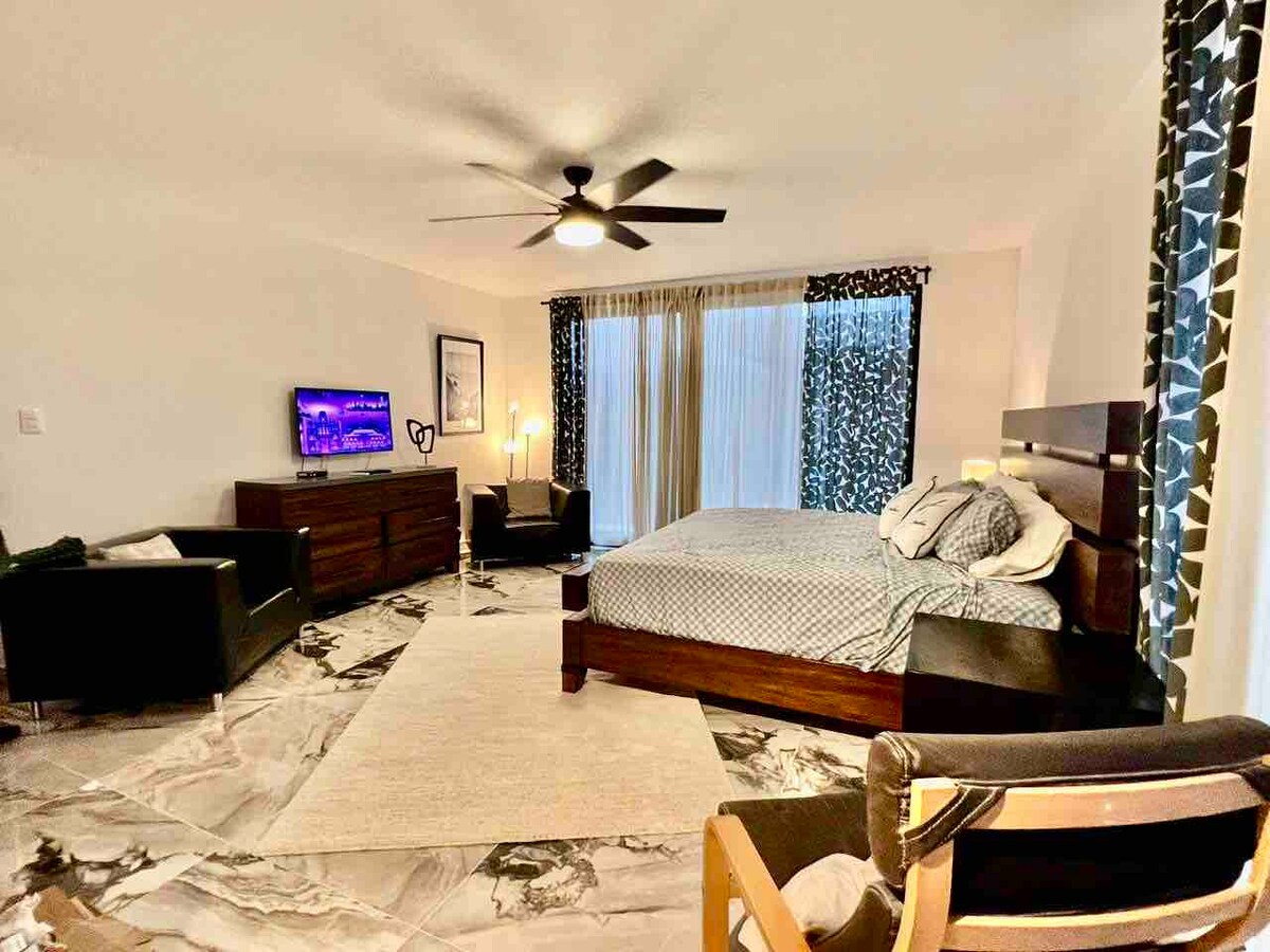A spacious bedroom is furnished with a large bed and dark wooden furnishings. A seating area with black chairs is present, along with a dresser. Large sliding glass doors allow natural light to fill the room, and a flat-screen TV is mounted on the wall.