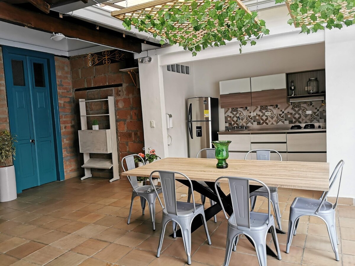 An open dining area features a large wooden table surrounded by six metal chairs. Natural light floods the space, highlighting the adjacent kitchen, equipped with modern appliances. A vibrant green plant decorates the table, and exposed brick adds textural contrast to the contemporary design.