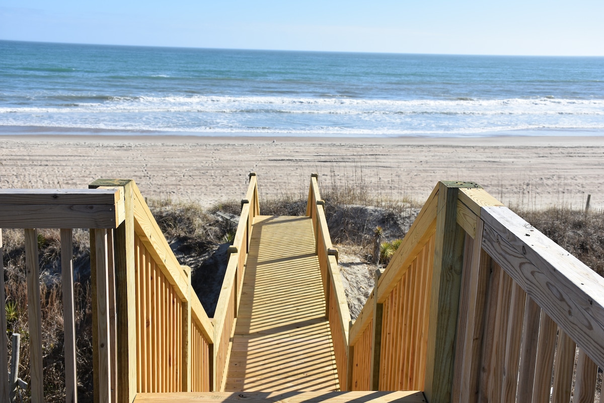 A wooden stairway leads from the cottage to the sandy beach, framed by coastal vegetation. The beach stretches into the distance, meeting the calm ocean under a clear blue sky. Gentle waves are visible, enhancing the tranquil setting.