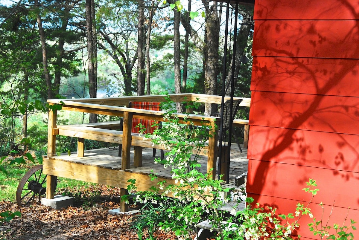 A wooden deck extends from the cottage, surrounded by trees and greenery. The deck features a plaid blanket draped over a chair, inviting relaxation while offering peaceful views of the natural surroundings. The vibrant red wall of the cottage adds a warm contrast to the landscape.