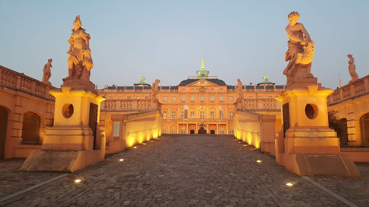 The image shows a historic palace building illuminated at dusk, flanked by elegantly carved statues. A cobblestone pathway leads towards the entrance, which is highlighted by warm lights. The architecture features ornate details and a prominent roof structure that adds to its grandeur.