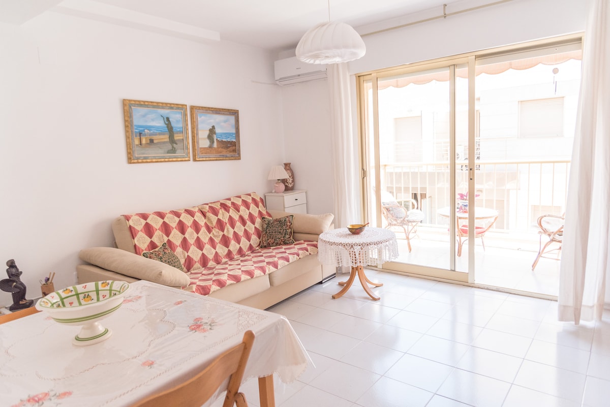 A light-filled living area features a comfortable beige sofa adorned with a patterned quilt. A small round table with a lace cover sits nearby, while a pair of framed artworks adorns the wall. Sliding glass doors lead to a balcony, allowing for natural light.