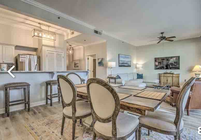 A spacious living area features light wood flooring and natural light from large windows. A dining table with decorative chairs sits adjacent to a fully equipped kitchen with stainless steel appliances. A comfortable seating area includes a sectional couch and a coffee table, with a wall-mounted television visible.
