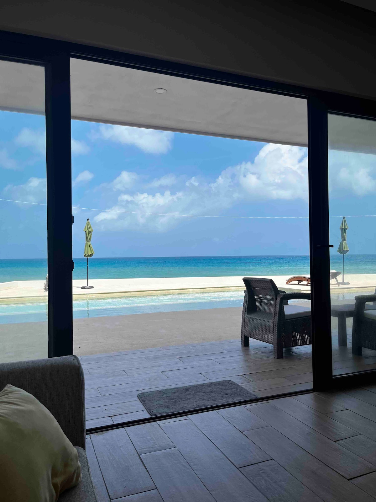 A spacious living area features large glass doors that open to a serene beachfront view. Umbrellas are positioned on the sand, overlooking the turquoise sea and a pool area, inviting relaxation under the sky.