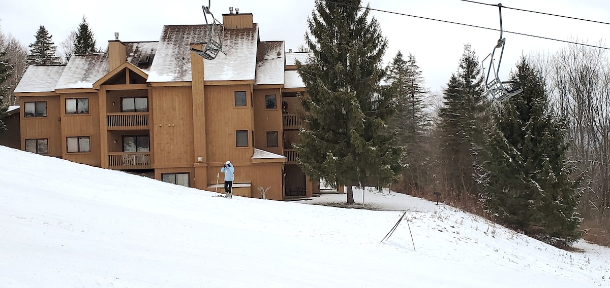 A cozy condo building is nestled among snow-covered trees. The structure features balconies and large windows, while a skier is seen navigating down the slope, showcasing the ski-in/ski-out access. The surrounding landscape is tranquil, with a blanketed layer of fresh snow.