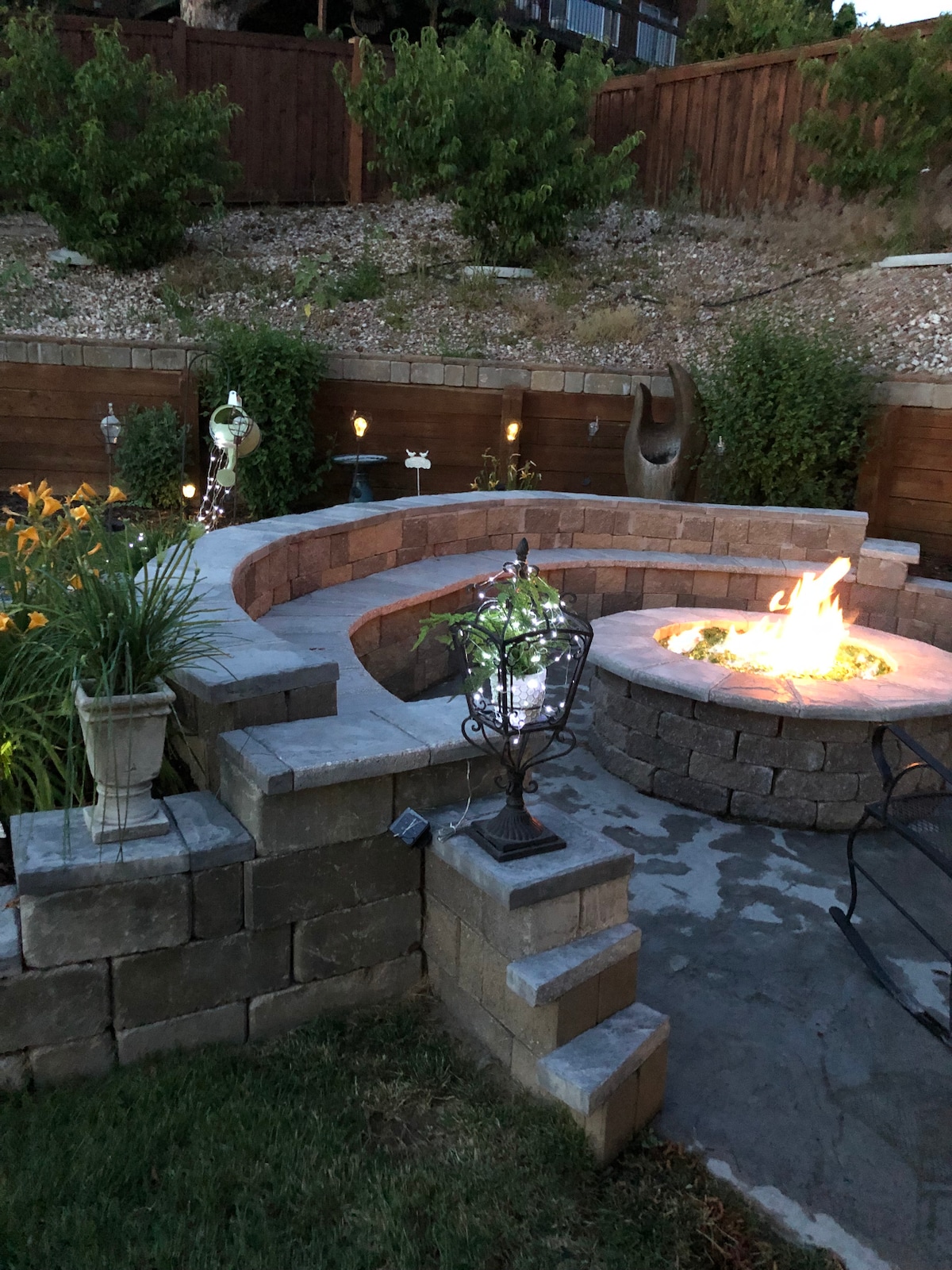 A cozy outdoor seating area is framed by a curved stone wall, surrounding a central fire pit. Lush foliage and flowering plants add greenery, while soft lighting provides a warm atmosphere. The area creates an inviting space for relaxation and gathering in the fenced backyard.