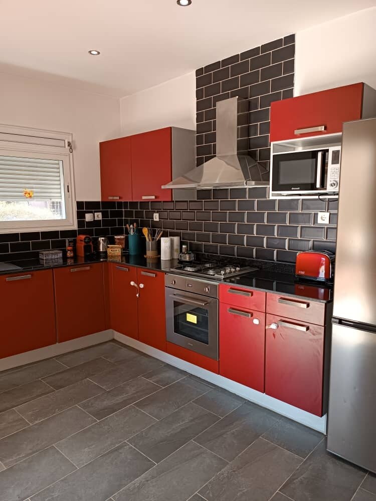 The modern kitchen features bold red cabinetry paired with dark tiled backsplash. Stainless steel appliances, including an oven and microwave, are integrated into the design. Counter space is ample, with various cooking utensils visibly arranged, ensuring a functional and welcoming cooking environment.