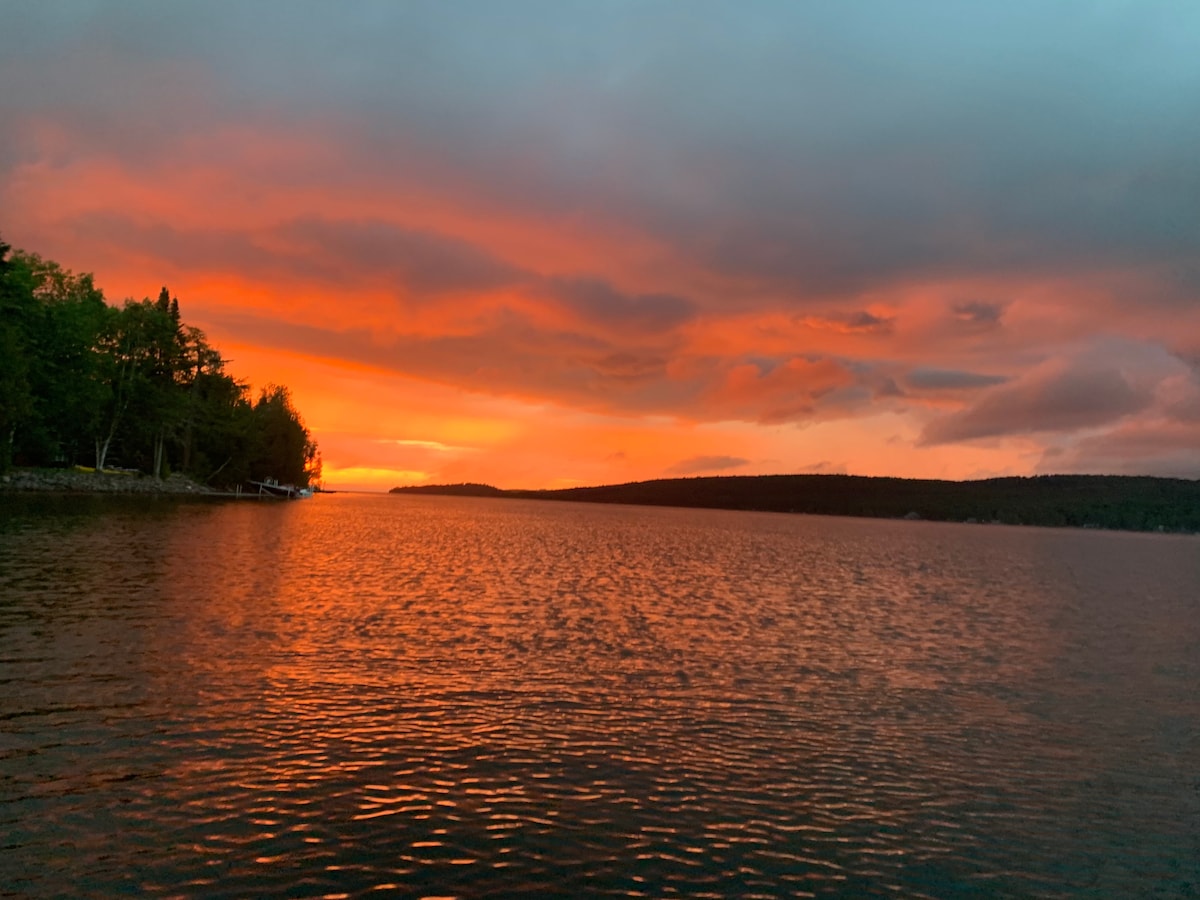 A serene lakeside view captures a vibrant sunset, with hues of orange and pink reflecting off the calm water. The shoreline is lined with trees, and silhouettes of distant hills can be seen under a cloudy sky, creating a peaceful atmosphere.