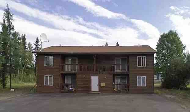 A two-story wooden structure is visible, featuring a symmetrical design with multiple windows on each level. A central entrance door is flanked by covered patios. Surrounding greenery and a clear sky contribute to the overall calm setting.