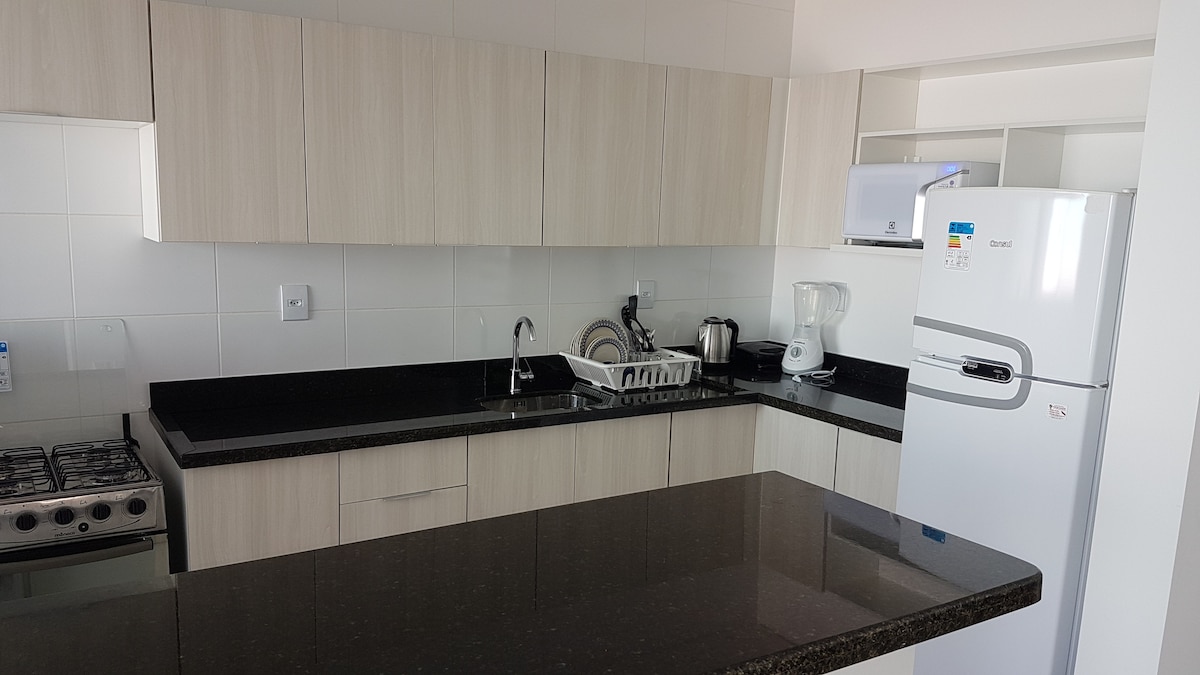 A modern kitchen is equipped with a black countertop, sleek cabinetry, and a gas stove. Essential appliances, including a refrigerator, kettle, and blender, are present. A sink and drying rack can also be seen, creating a functional cooking space.