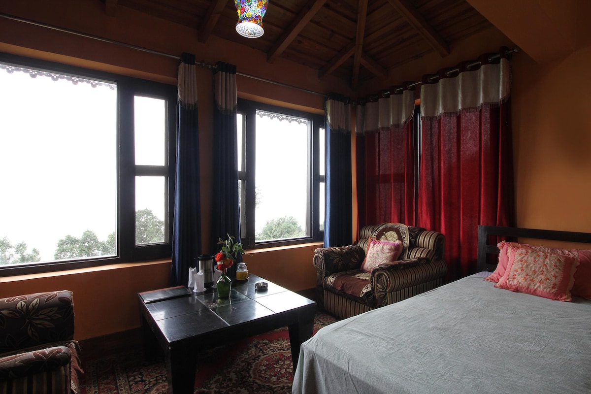 A cozy room features a spacious bed and two comfortable armchairs. Natural light floods in through large windows, adorned with elegant curtains. A dark wooden table sits in the center, accompanied by a vase of flowers, enhancing the inviting atmosphere of the space.