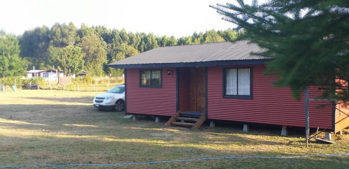 A red cabin is presented with a welcoming entrance, featuring a front porch and a set of steps. A white vehicle is parked nearby on the grassy area. Lush greenery and trees surround the cabin, creating a peaceful environment.