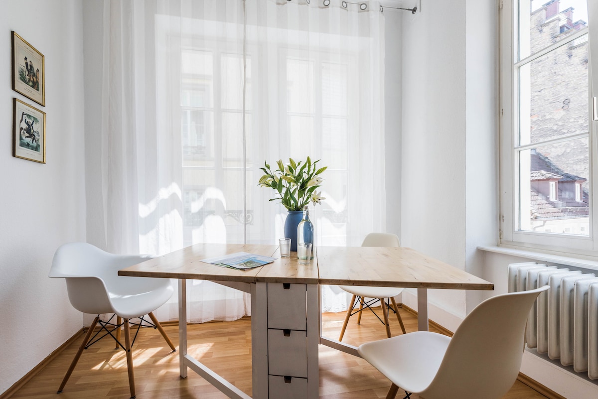 A bright dining area features a wooden table at its center, surrounded by three modern chairs. Natural light filters through sheer curtains, highlighting the minimalist decor. A vase with fresh flowers adds a welcoming touch to the space.
