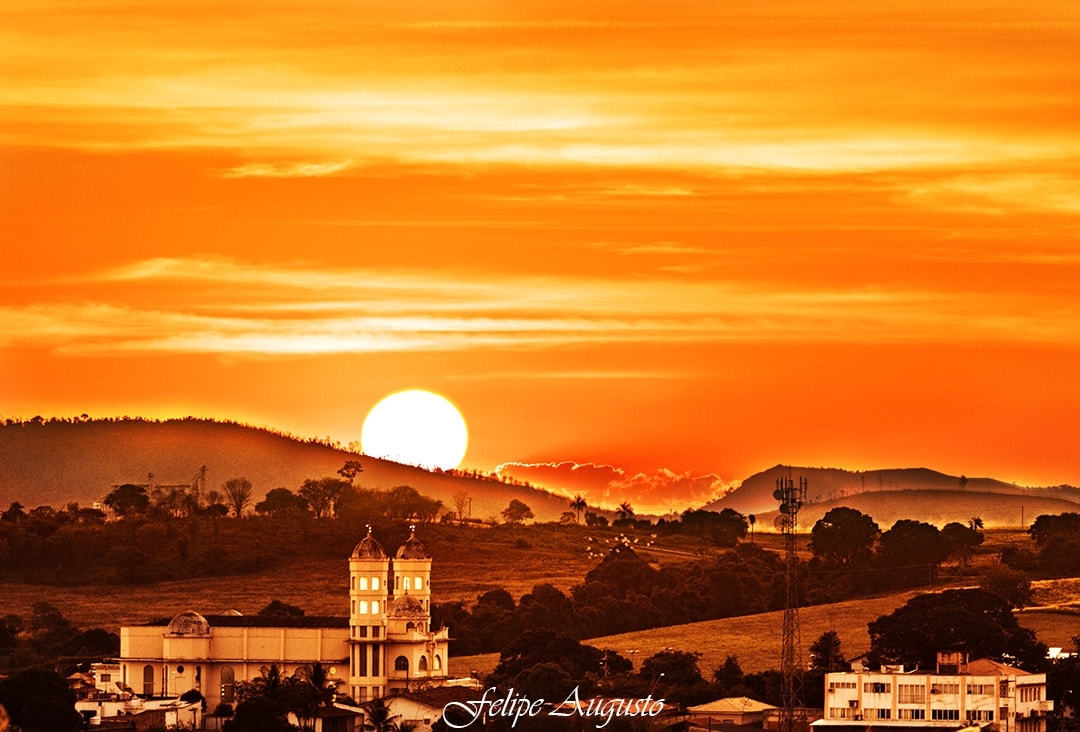 A sunset is captured over a scenic landscape, with warm orange hues illuminating the sky. The sun is setting behind distant hills, and the silhouette of a tower can be seen in the foreground, adding depth to the tranquil scene.