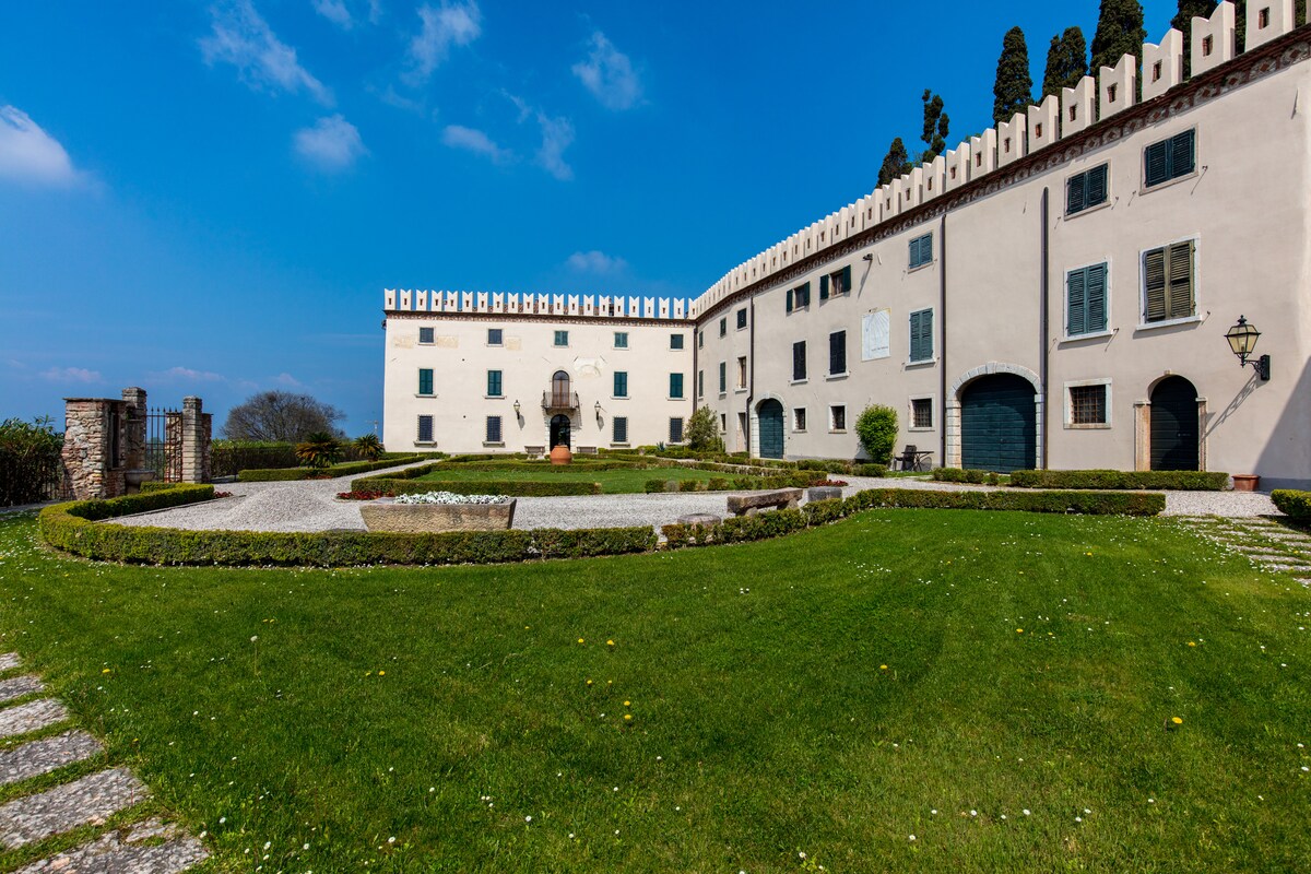 The exterior of the historic villa is showcased, featuring a manicured lawn bordered by hedges and flowerbeds. The building's architecture includes tall, arched windows and a decorative parapet. A stone pathway leads through the landscape, with a statue positioned in the central garden area.