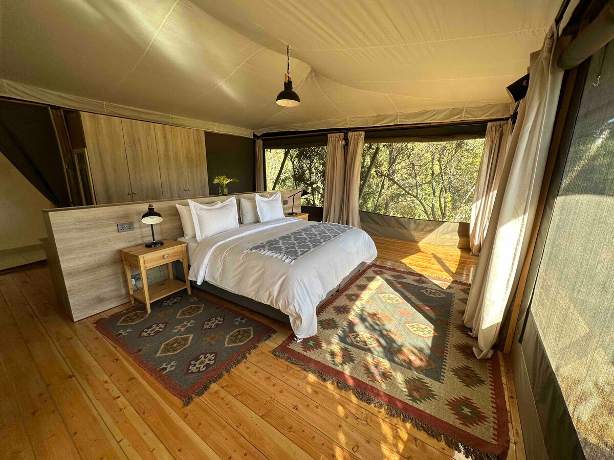 The bedroom area features a spacious bed with white linens, accompanied by a bedside table. Natural light filters through large windows, revealing views of the surrounding nature. The wooden floor is complemented by patterned rugs, enhancing the warm and inviting ambiance.