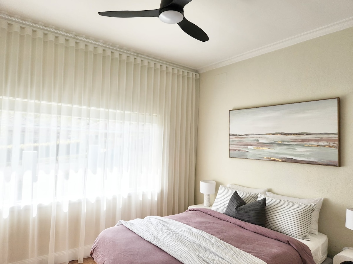 A spacious bedroom features a large bed adorned with soft bedding and decorative pillows. Natural light filters through sheer curtains, illuminating the room. A modern ceiling fan is positioned above, and stylish bedside lamps provide additional lighting. A landscape painting hangs on the wall.