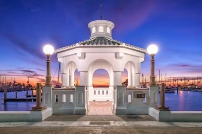 A bright waterfront gazebo with white architecture stands against a vibrant sunset. The structure features arched openings and a tiled roof, illuminated by elegant lamps. Sailboats can be seen in the background, adding to the serene coastal ambiance.