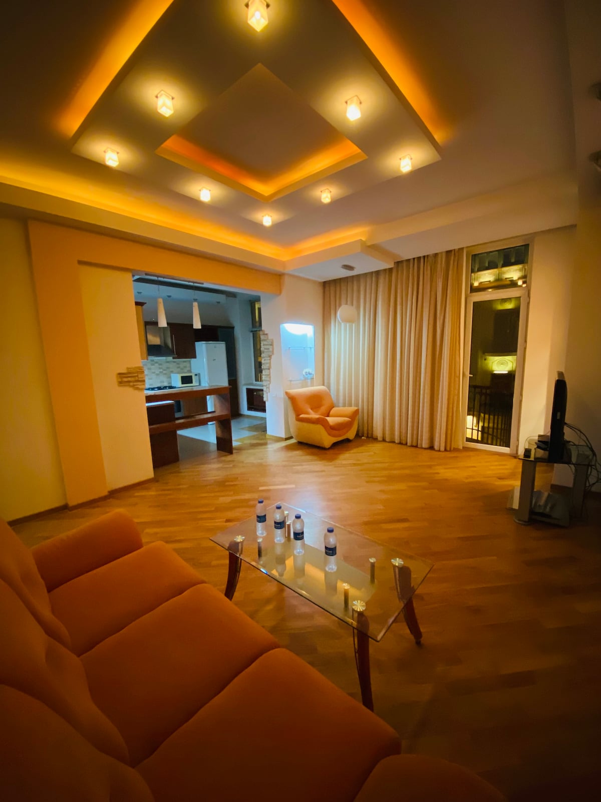 A living area is highlighted by warm lighting, featuring a comfortable orange sofa and a glass coffee table. Light wood flooring complements the space, while large windows with light curtains offer views of the surroundings. An inviting armchair adds to the seating options.