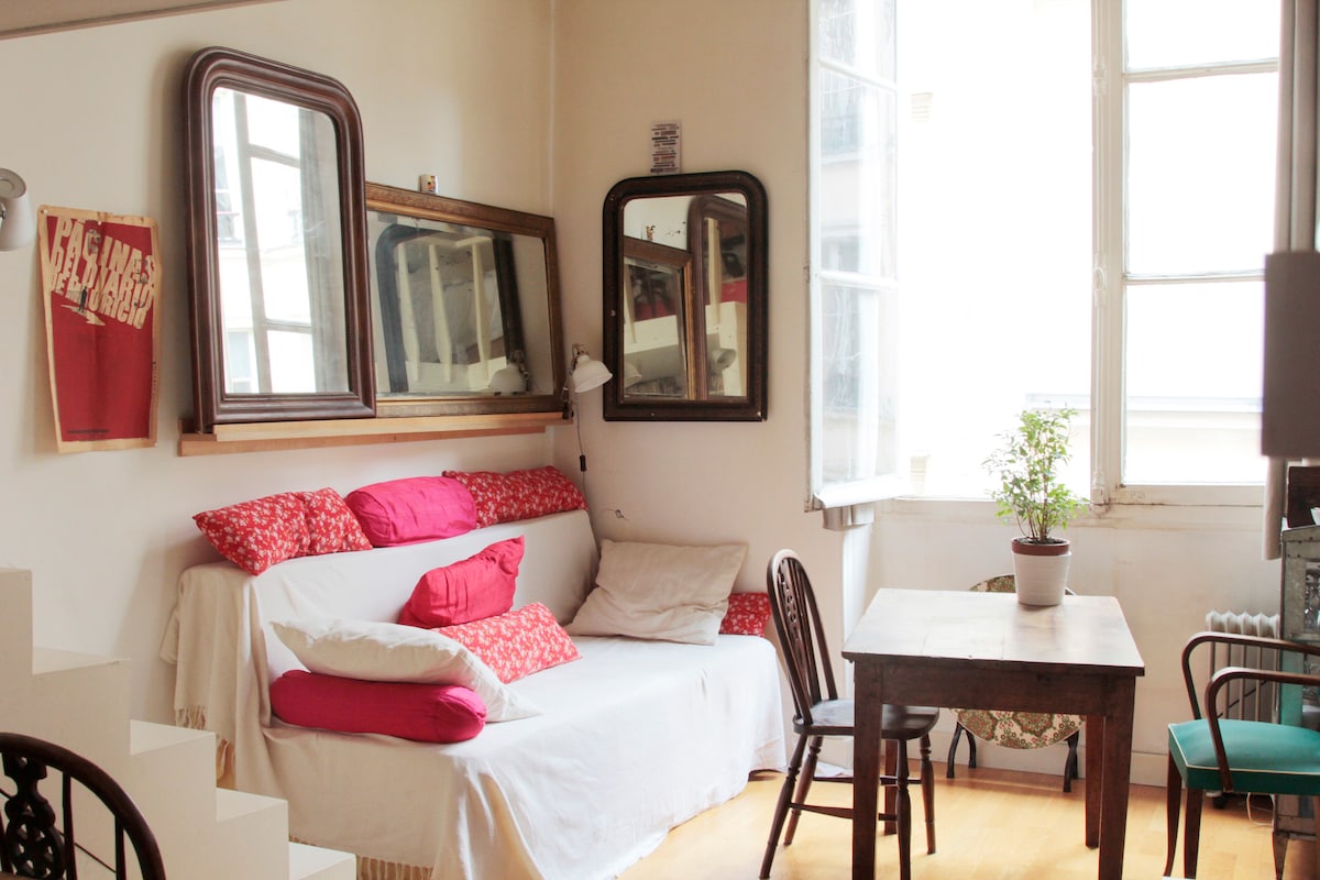 A comfortable seating area features a white sofa adorned with colorful cushions. Multiple mirrors reflect light in the space. A small wooden table and chair sit nearby, with a potted plant adding a touch of greenery. Large windows allow ample natural light.