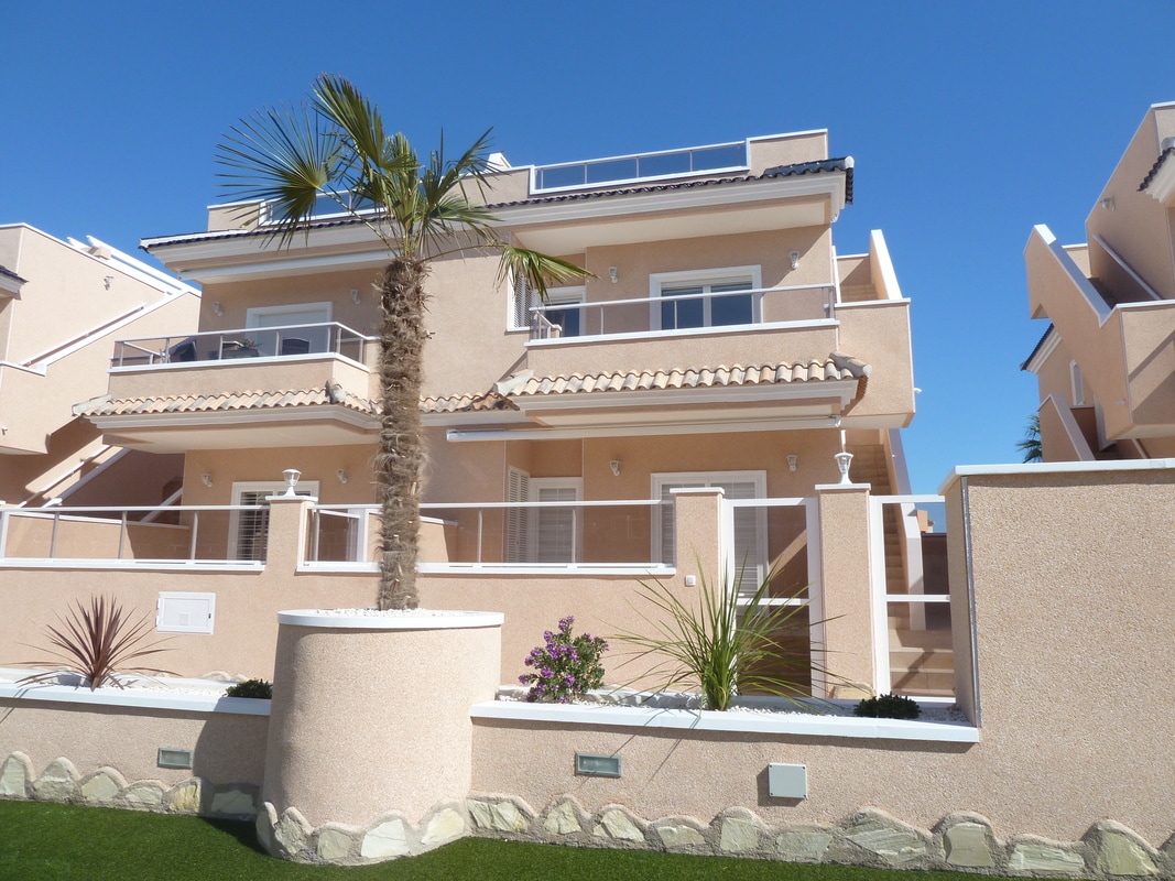 A modern duplex building with two levels is showcased, featuring light-colored exterior walls and tiled roofing. Lush greenery, including a palm tree and ornamental plants, adds to the well-maintained surroundings. A clear blue sky serves as the backdrop, emphasizing the welcoming atmosphere of the property.