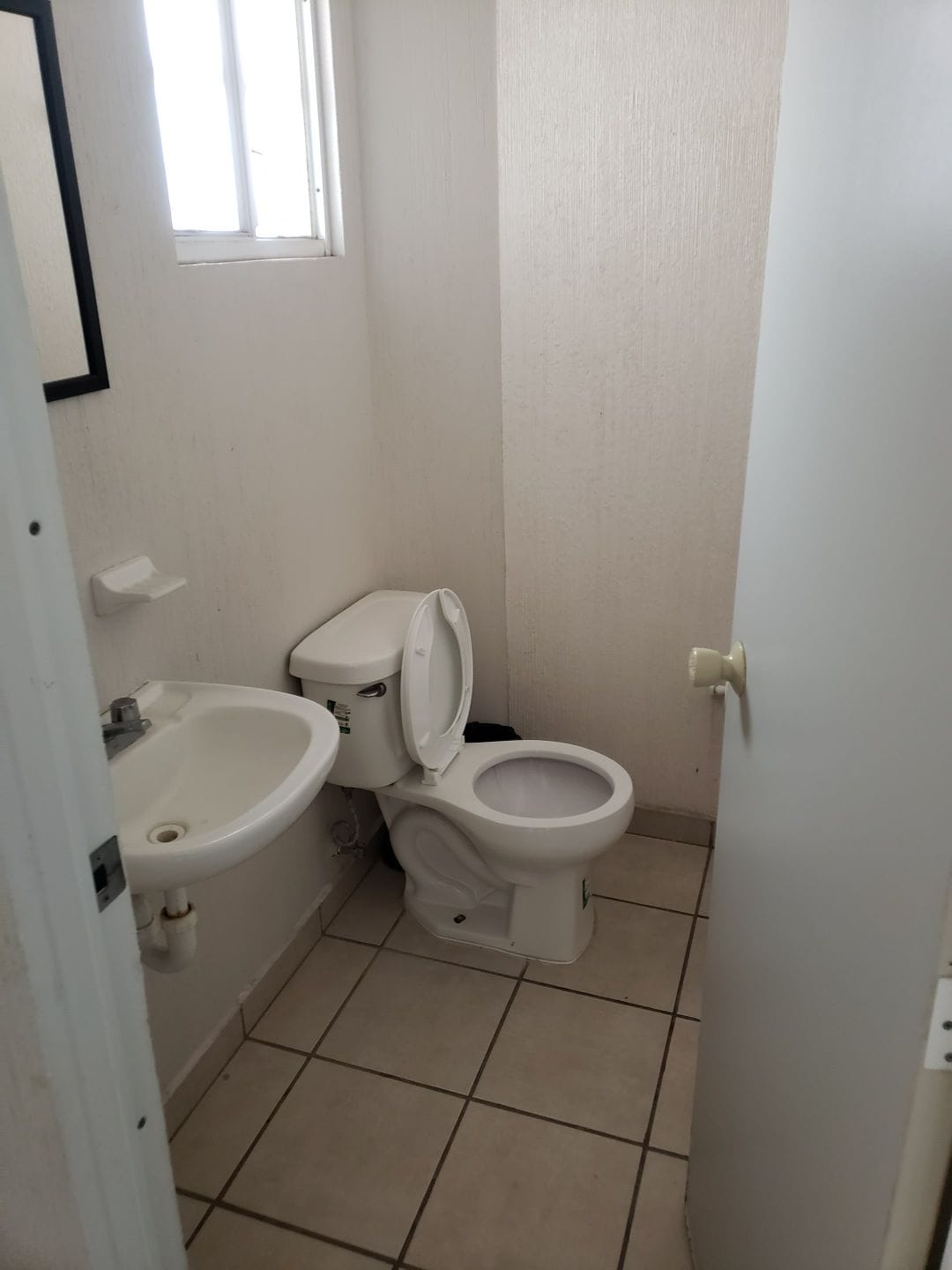 A compact bathroom is depicted with a toilet and a small sink installed on the wall. A window allows natural light to enter, and light-colored tiles cover the floor. A mirror above the sink completes the functional space.