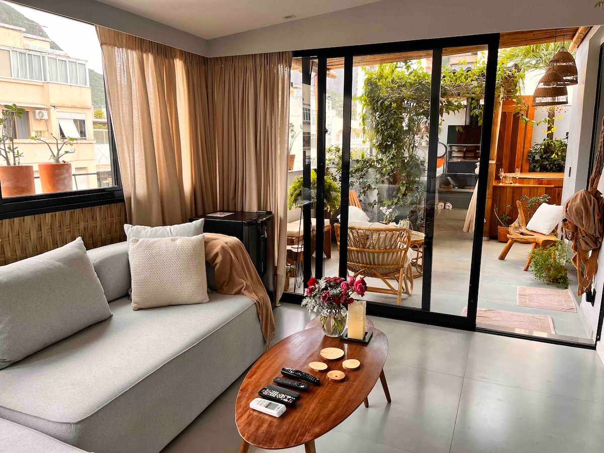 A bright living area features a comfortable gray sofa, accentuated by light-colored pillows. A wooden coffee table holds remote controls and decorative items. Large glass doors lead to a vibrant outdoor space filled with greenery, integrating nature into the living environment.