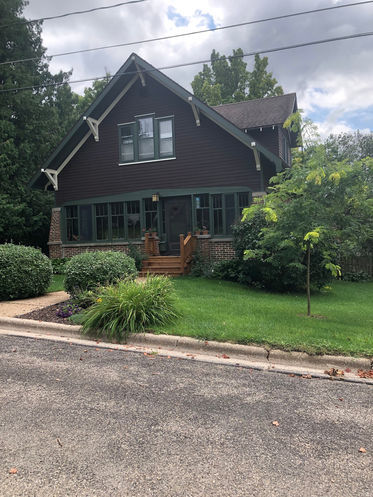 The exterior of a charming 1920's Arts and Crafts home is depicted, featuring dark wood siding and classic architectural details. A welcoming front porch with steps leads to the entrance. Surrounding greenery includes a well-maintained lawn and shrubs, contributing to the home's inviting presence.