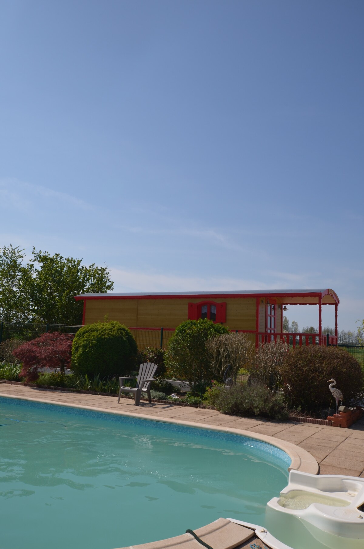 A vibrant roulotte is set against a clear blue sky, surrounded by lush greenery and colorful landscaping. A shimmering pool is visible in the foreground, accompanied by a sun lounger positioned nearby. The scene reflects a serene outdoor environment, ideal for relaxation.