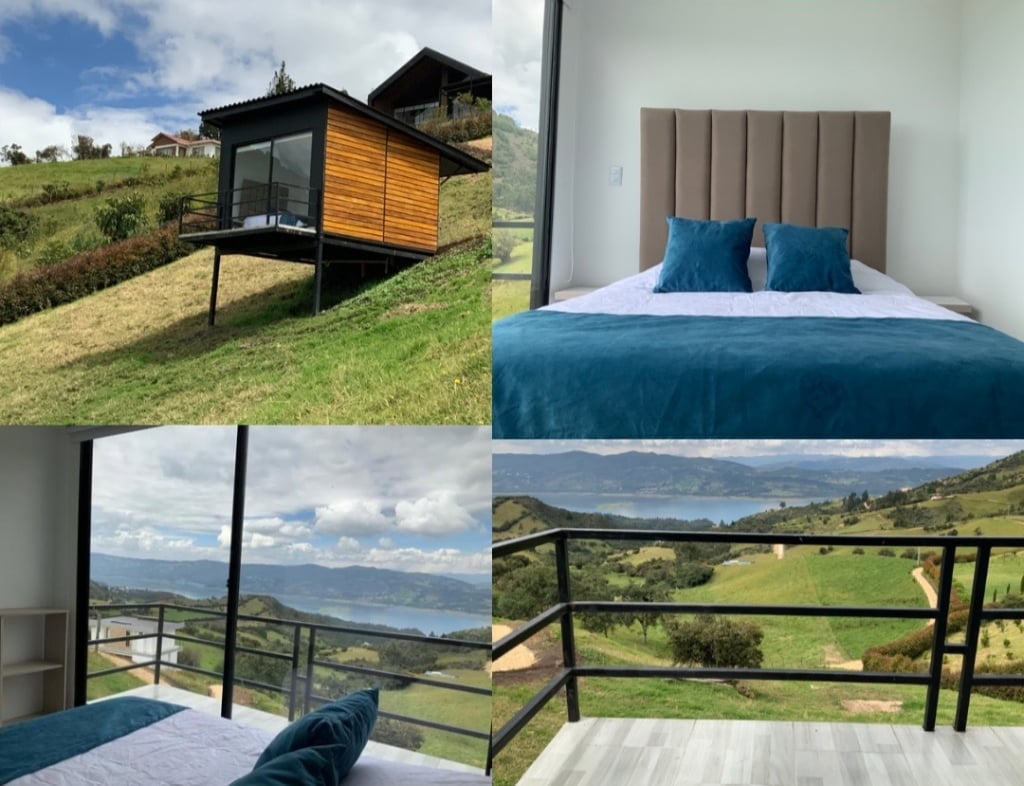 A modern cabin is situated on a grassy hillside, featuring large windows offering panoramic views. The cozy bedroom is furnished with a spacious bed adorned with soft blue bedding. Natural light fills the space, highlighting the tranquil landscape and the nearby water reservoir.