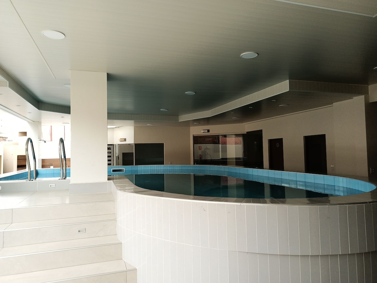 An indoor pool area features sleek, rounded edges with light-colored tiles. Steps lead into the water, and large windows provide natural light. The surrounding space offers a modern, open layout, enhancing the serene environment.