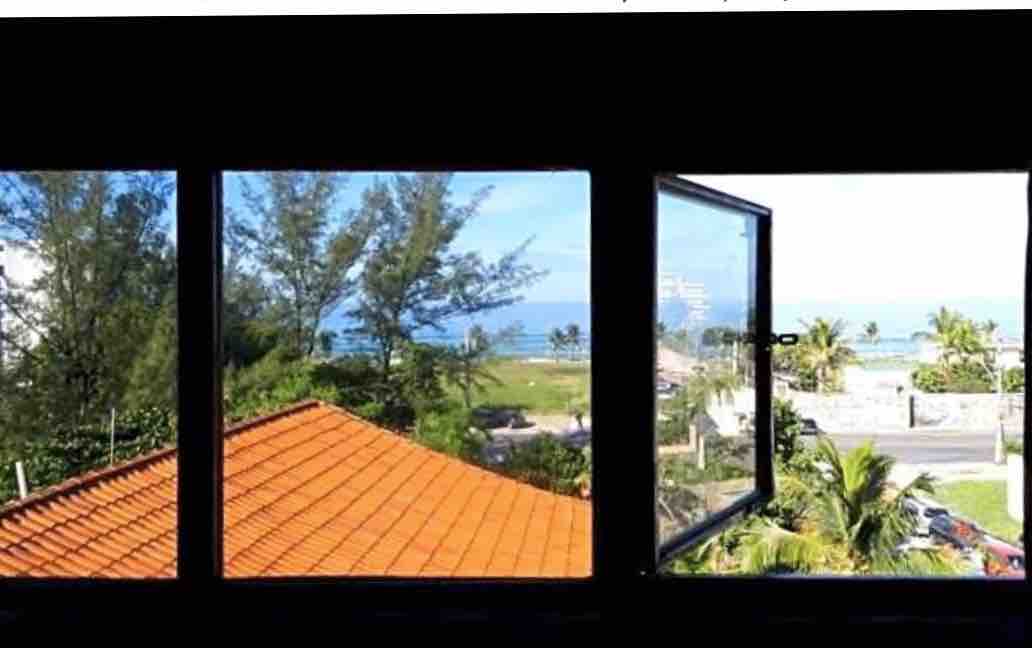 A panoramic view is captured through three large windows, showcasing lush greenery and the ocean in the distance. The vibrant orange roof tiles can be seen in the foreground, complemented by palm trees and open sky.