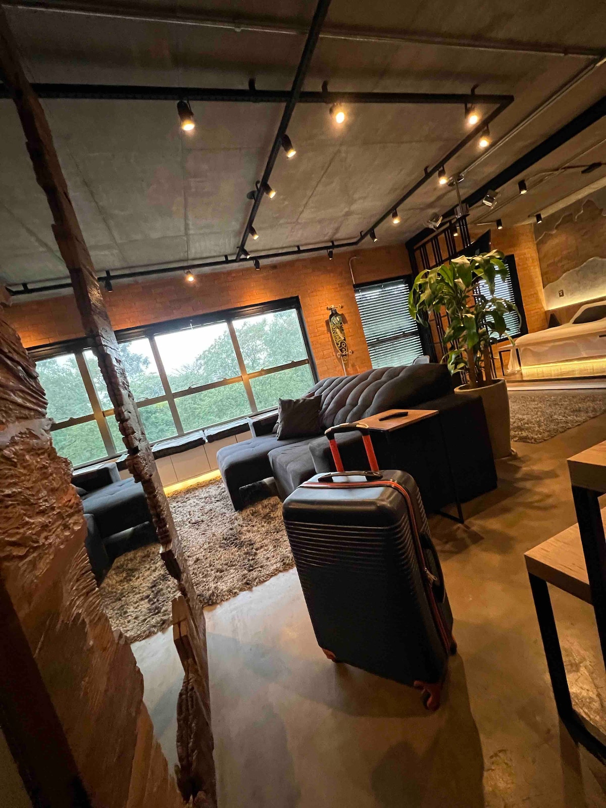 An inviting living area is showcased, featuring a comfortable dark sofa and a textured area rug underfoot. A suitcase is positioned nearby, and large windows provide natural light, framed by industrial design elements. Greenery adds a touch of nature to the space.