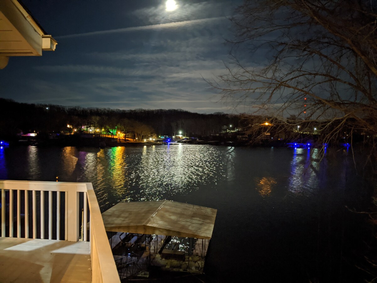 A night view of the lake features shimmering water illuminated by moonlight, reflecting various colors from nearby lights. The peaceful setting includes a covered dock, framed by trees and distant shorelines, creating a serene outdoor atmosphere.