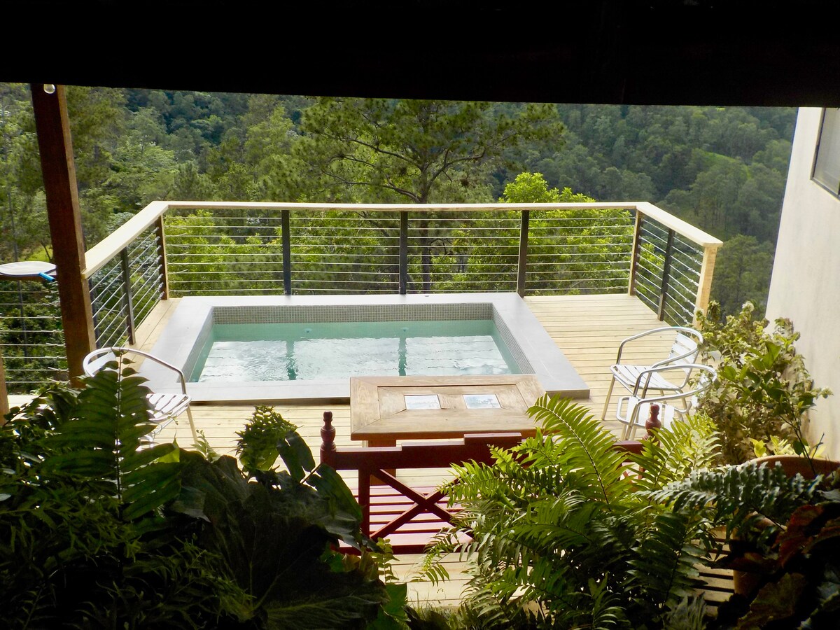 A wooden deck features a private jacuzzi surrounded by lush greenery. Two chairs are positioned nearby, offering a relaxing spot to enjoy views of the mountainous landscape. The jacuzzi area is illuminated by natural light, enhancing the tranquil ambiance.