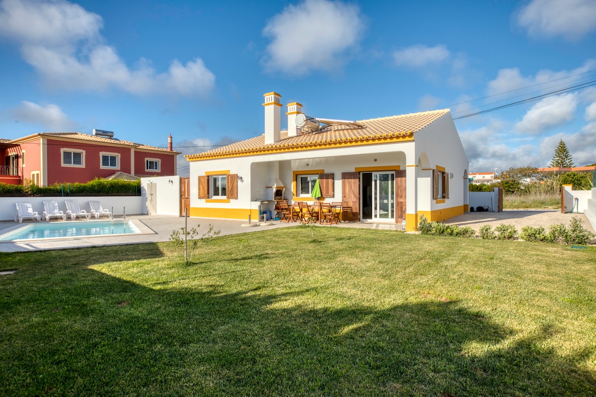 A spacious house is depicted with a well-maintained garden and a swimming pool. Sunlight highlights the clean lines of the exterior, featuring warm hues. The patio area includes a dining set with chairs, providing an ideal space for outdoor meals. Lush grass surrounds the amenities.
