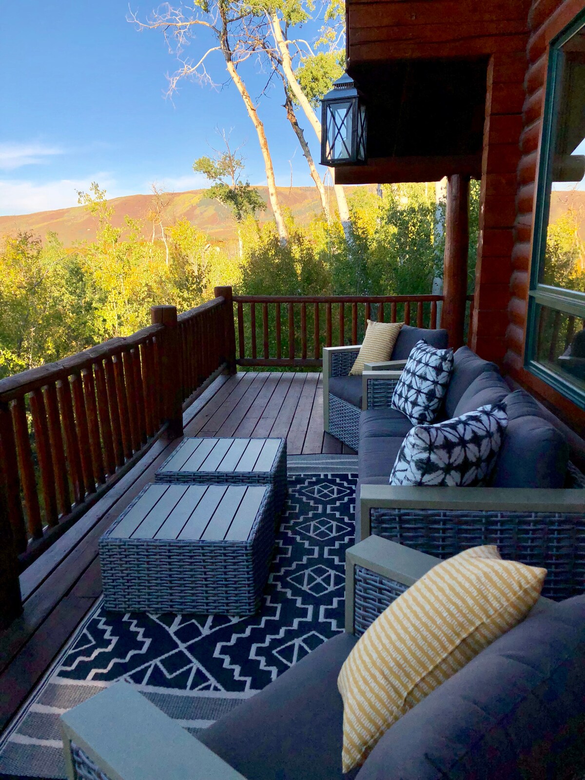 A cozy outdoor seating area is showcased on a deck, featuring a set of gray wicker sofas adorned with patterned cushions. Two side tables are positioned on a decorative rug, with views of the surrounding trees and mountains in the background.