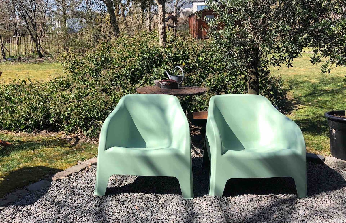 Two light green chairs are positioned in a serene garden setting, surrounded by greenery. A small round table sits nearby, complementing the outdoor space. Sunlight filters through the trees, creating a peaceful atmosphere perfect for relaxation.
