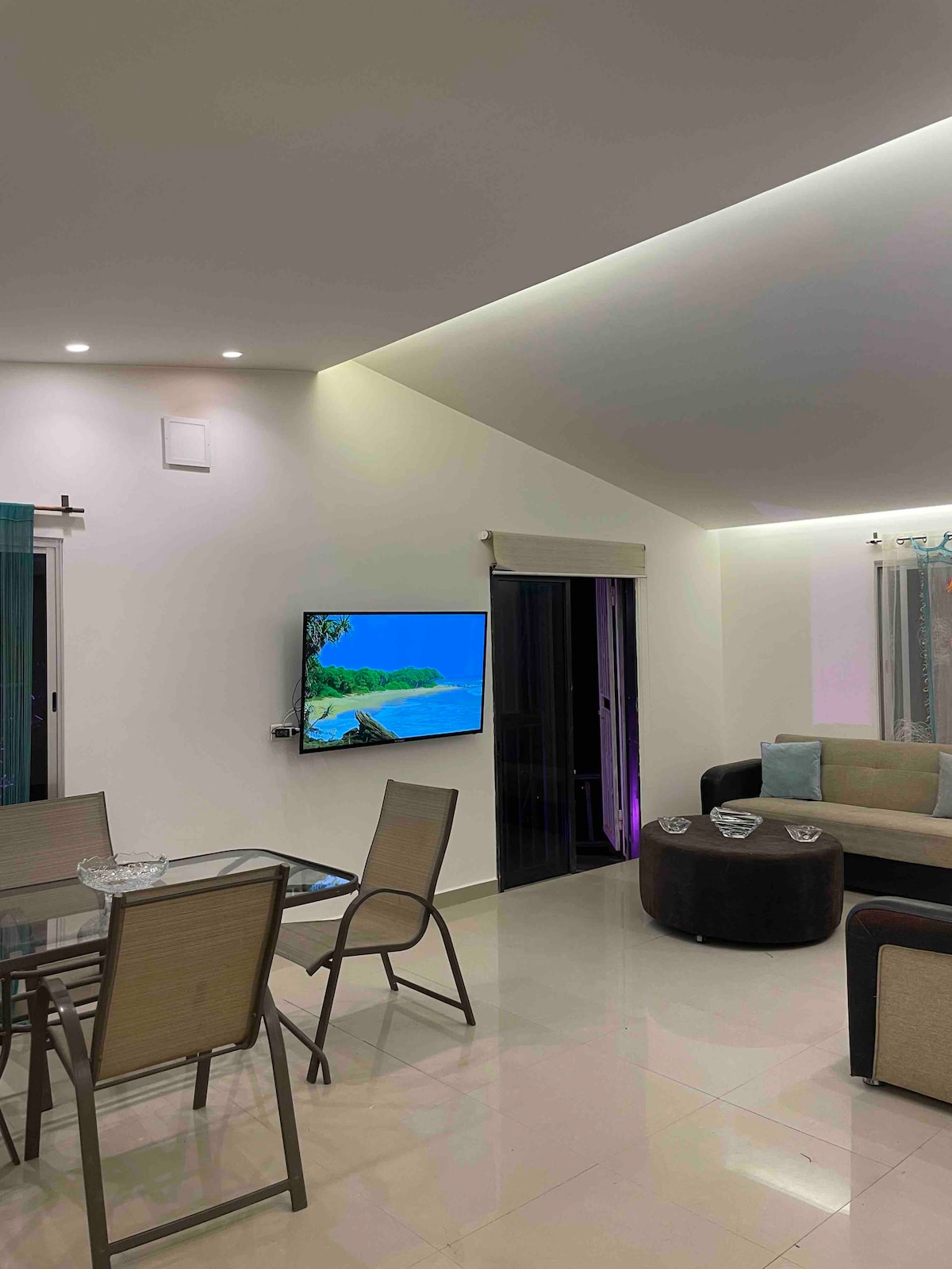 An open living area is visible, featuring a smart TV mounted on the wall. A glass dining table with four chairs is positioned adjacent to a comfortable seating arrangement, which includes a dark round ottoman and a light-colored sofa. Natural light enters through large sliding doors.