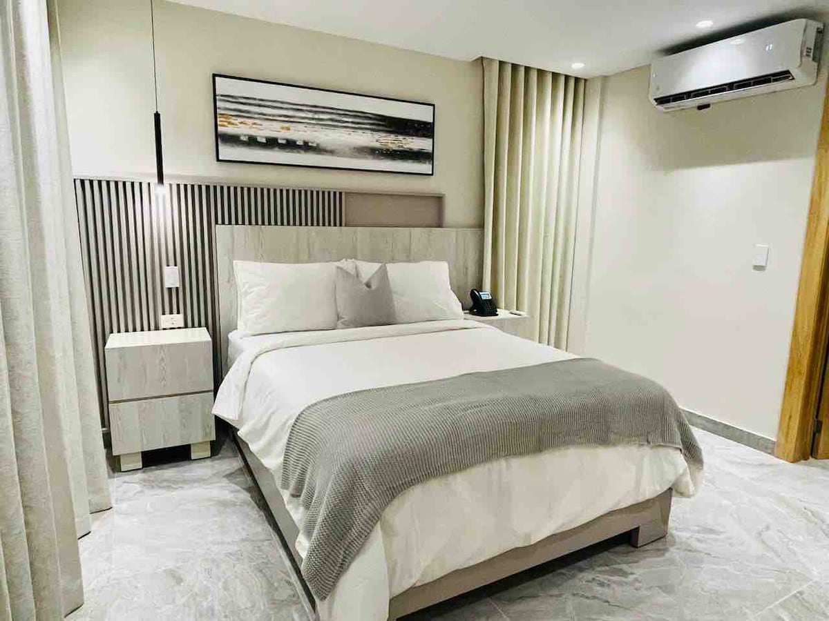 A sleeping area features a spacious bed with neutral bedding and a light gray throw. Minimalist nightstands are positioned beside the bed, and soft curtains frame the window. Modern artwork hangs above the headboard, while air conditioning is visible on the wall.