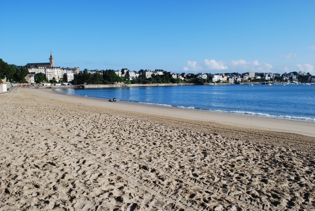 A sandy beach is visible with clear blue skies above. Gentle waves lap the shoreline, creating a serene atmosphere. In the distance, a coastal town with several buildings and a steeple can be seen along the shoreline. The beach appears spacious and inviting.