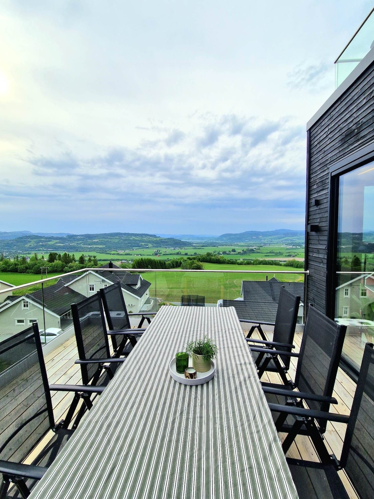 A spacious outdoor dining area is presented on a deck, featuring a long table surrounded by black chairs. The landscape beyond showcases rolling hills and valleys, complemented by a serene sky, creating a tranquil setting for meals or relaxation.