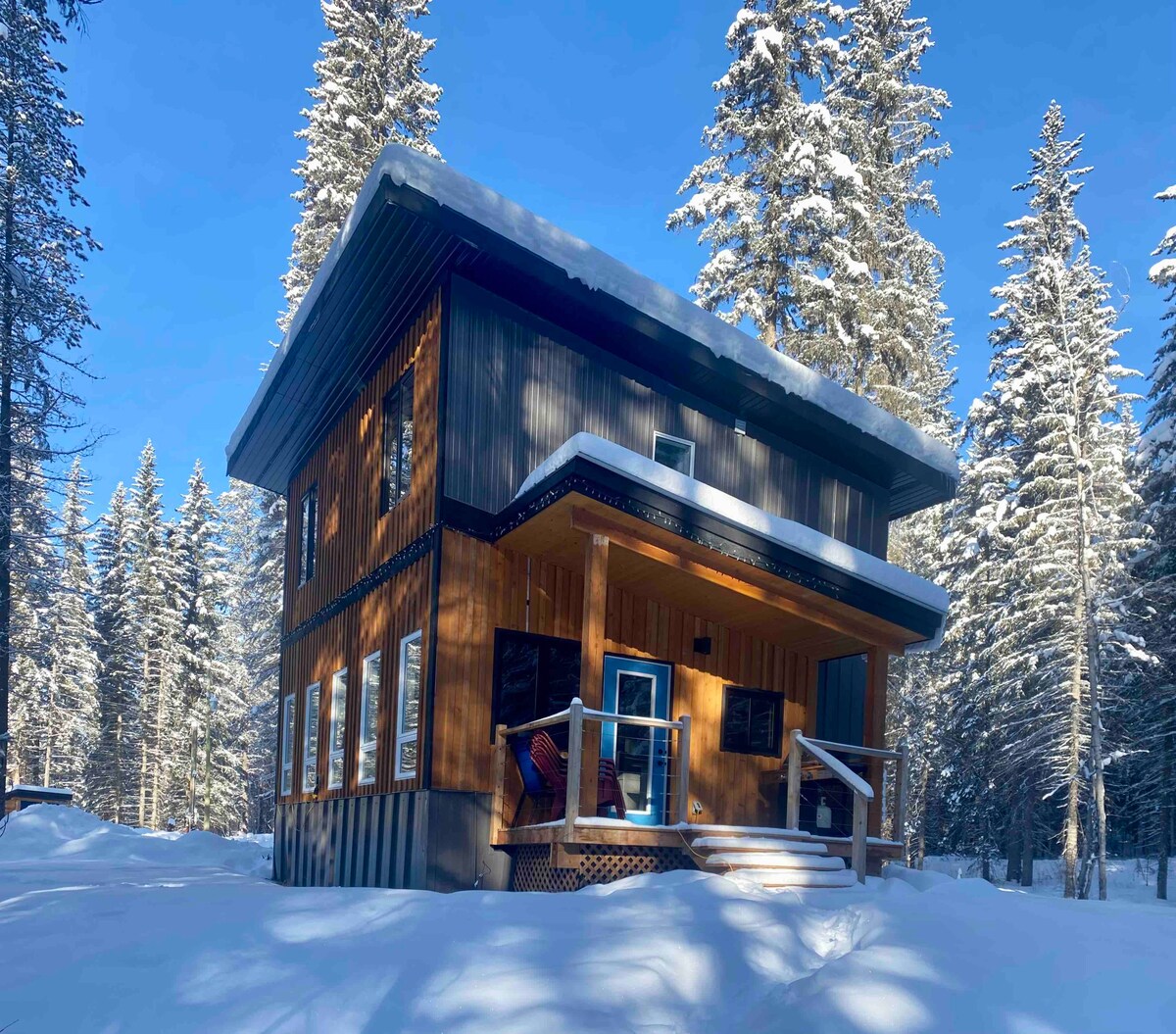 The two-story chalet is situated amidst a snowy forest setting, featuring a wooden exterior with large windows. Snow covers the ground, and tall trees rise in the background, creating a serene environment. The entrance includes a covered porch with steps leading to the front door.