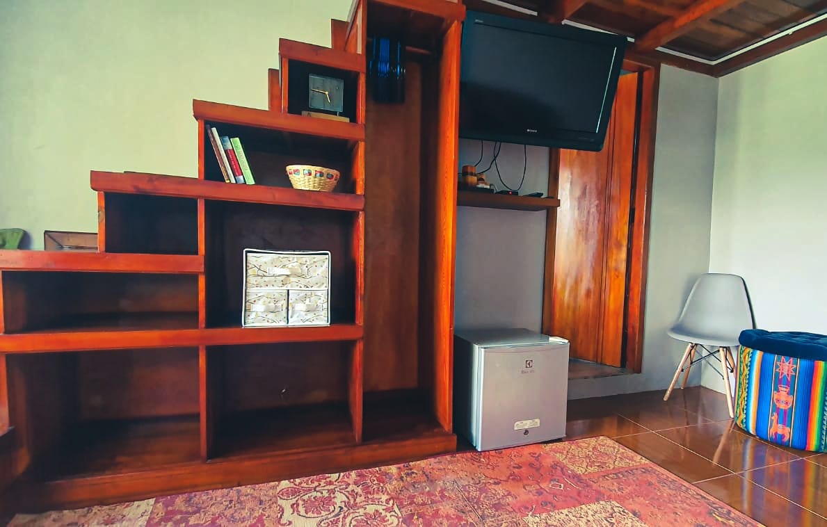 A wooden staircase with multiple shelves is featured, showcasing books and decorative items. A mounted television is positioned on the wall above a small cabinet, which is positioned next to a mini-fridge. A colorful ottoman adds a pop of design to the space.